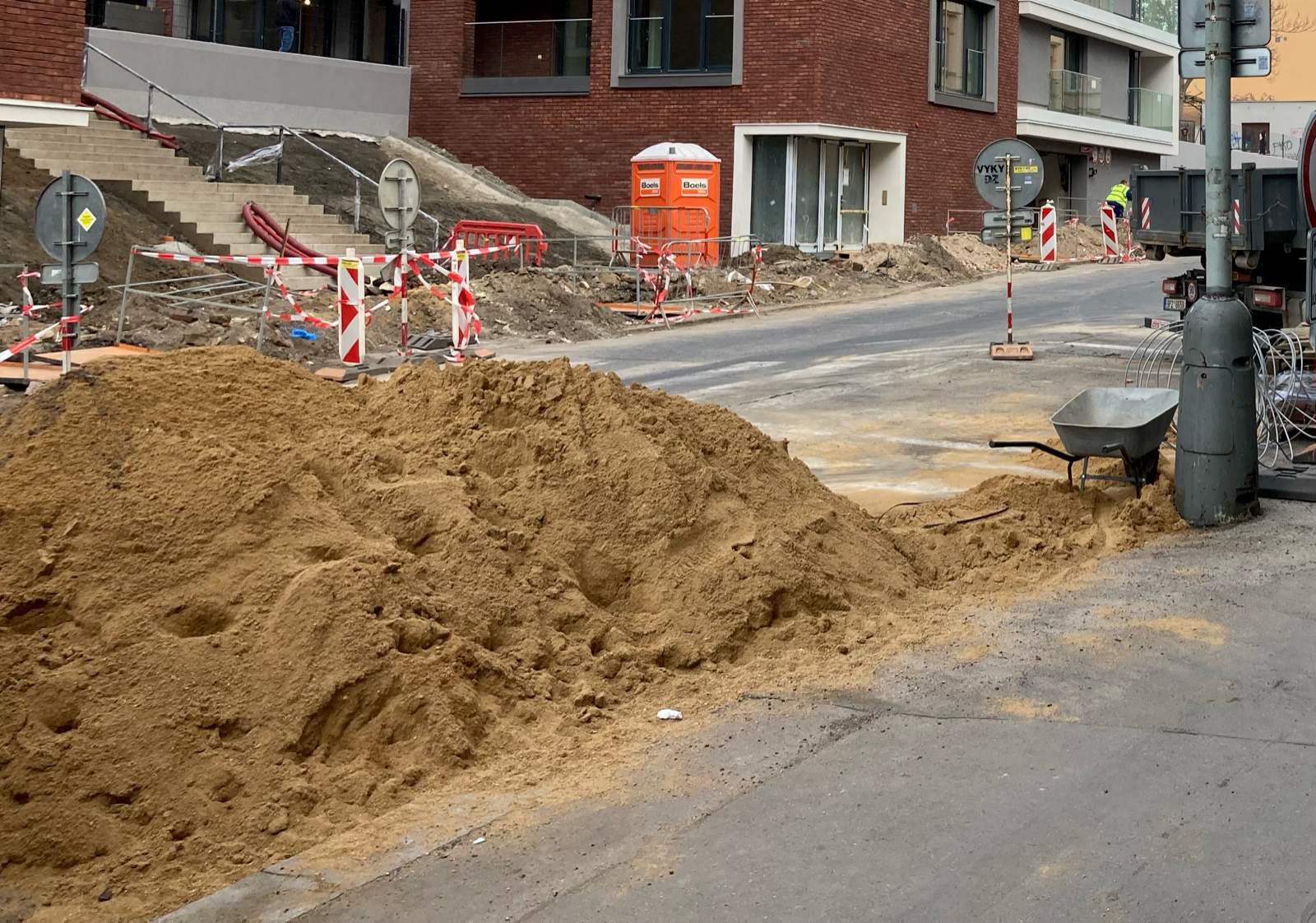 Montón de arena amarilla descargado en la calzada junto a una obra urbana, con vallas de obra y una carretilla al fondo