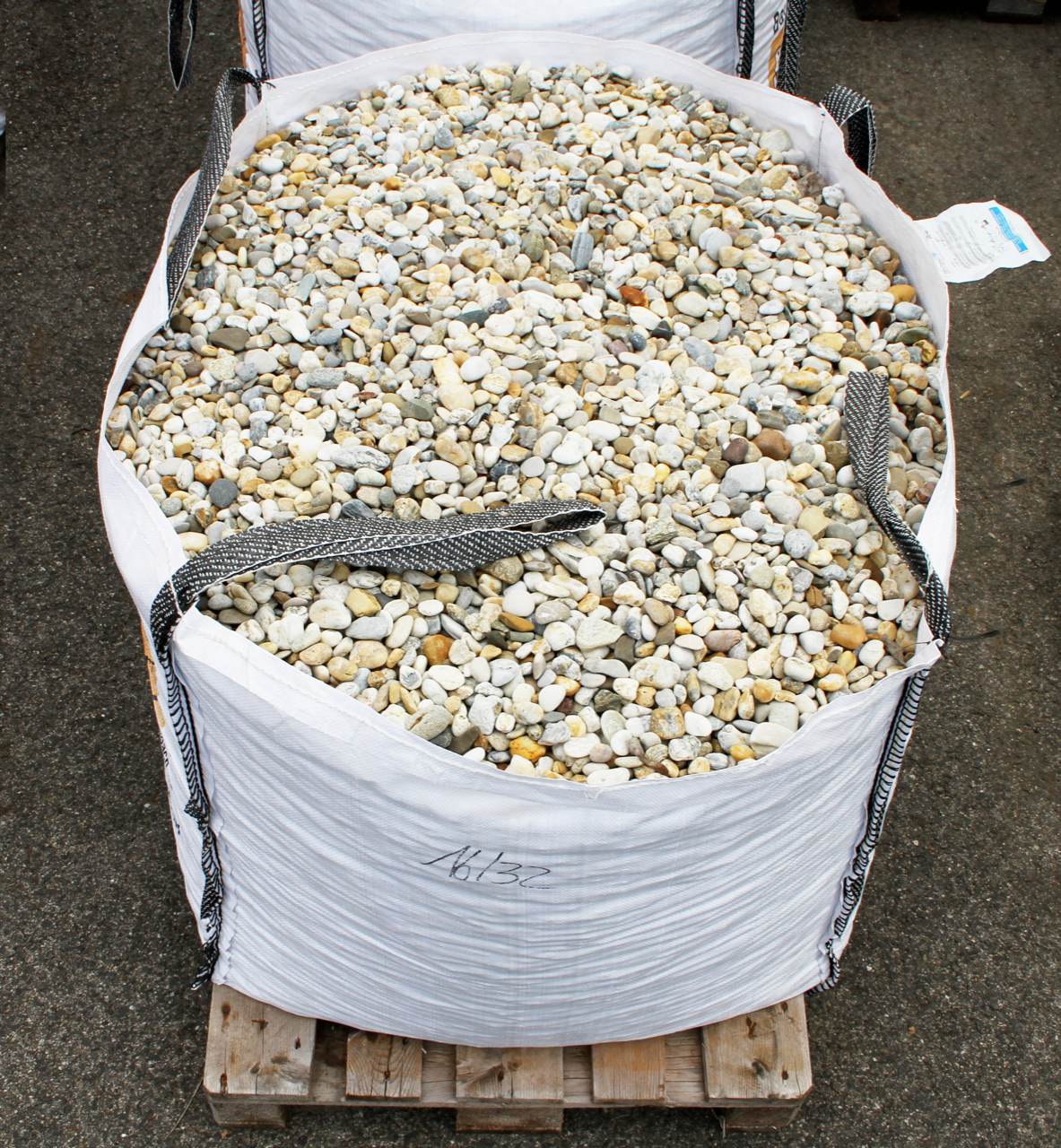 Big bag blanco lleno de gravilla redondeada, con las asas a la vista, sobre un palé de madera en el asfalto