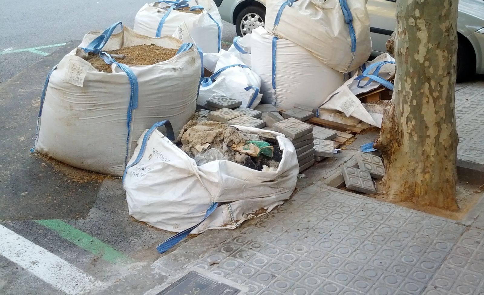 Varios big bags llenos de escombro y baldosas viejas en una acera de Barcelona, junto al alcorque de un árbol y coches aparcados