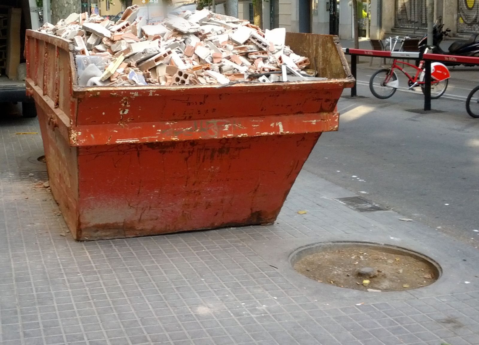 Contenedor metálico de obra, rojo y oxidado, lleno de cascotes de ladrillo hueco en una acera de Barcelona