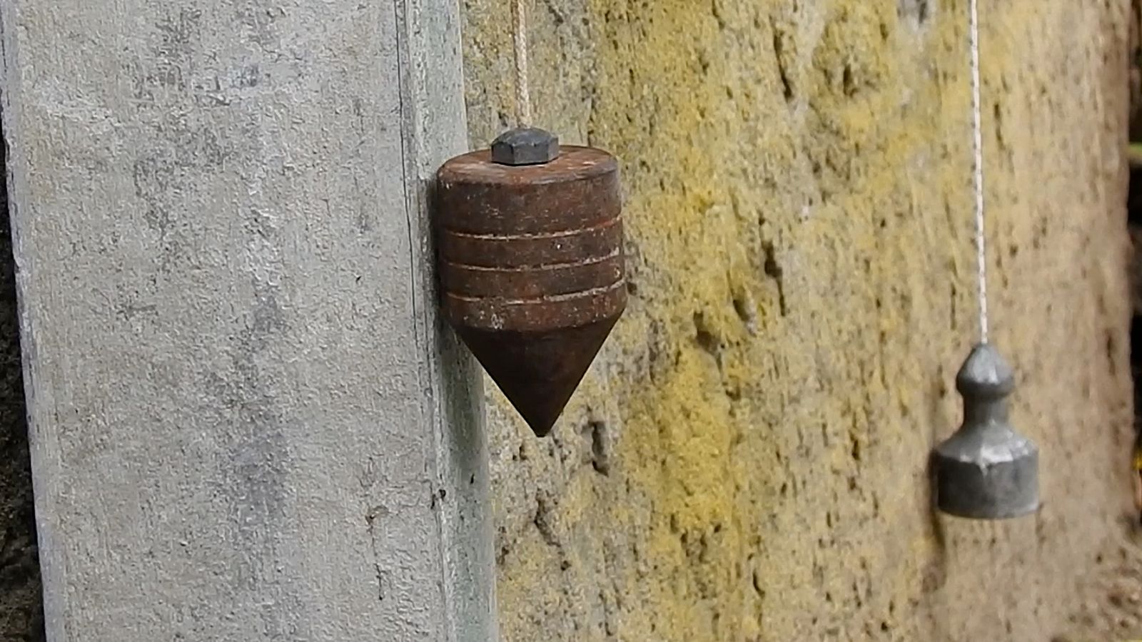Dos plomadas colgadas de su cordel junto a una pared, una de punta cónica y otra cilíndrica de albañil