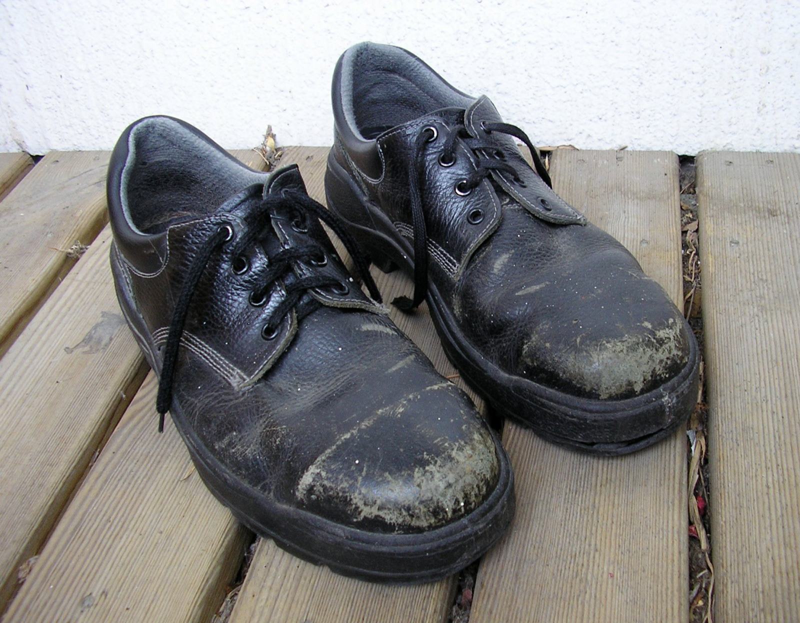 Par de zapatos de seguridad de piel negra, gastados y con manchas secas en la puntera, sobre un suelo de tablas